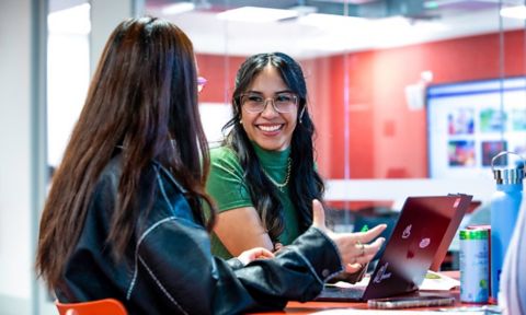 Two students on laptops, one facing away, one smiling at the other.