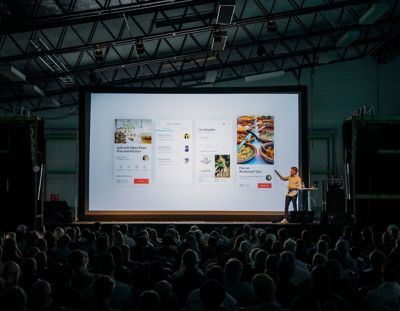 Presenter on stage talking and pointing to large screen in front of audience.