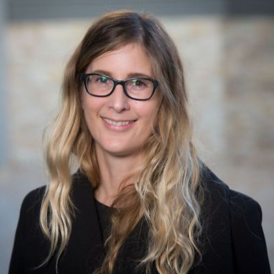 Louise Byrne faces forward and is smiling. Background is out of focus. Louise is wearing a black top and glasses.