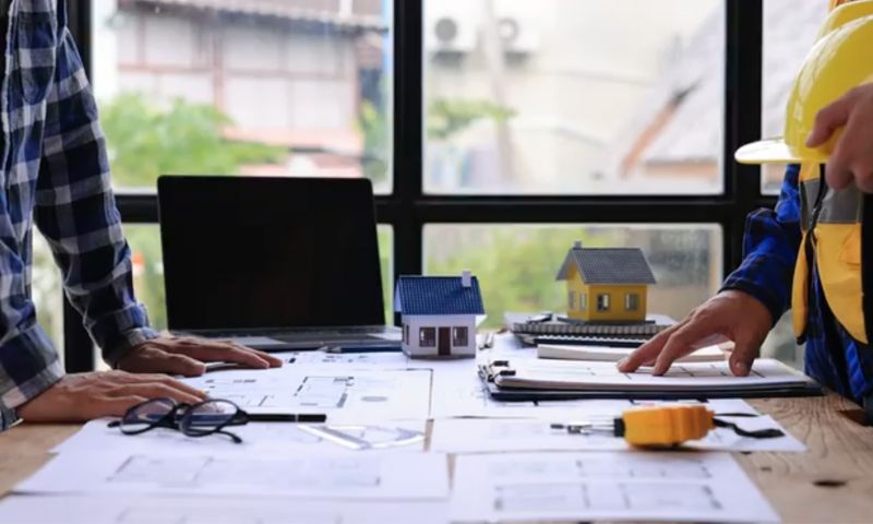 Two people facing each other across a table covered with plans.