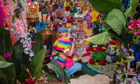 Artist sits among lots of colourful objects and fabrics in a big jumble, including a yellow birdcase, green leaves, a yellow dog toy, pink, blue, purple and green fabrics and fairy lights. 