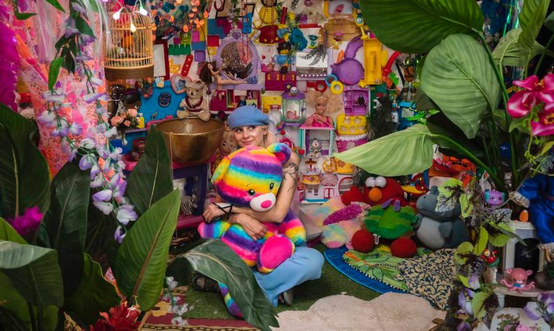 Artist sitting amongst lots of colourful objects and fabrics in a big jumble, including a yellow birdcase, green leaves, a yellow dog toy, pink, blue, purple and green fabrics and fairy lights. 