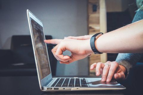 Photo of a laptop with a person operating the mouse. Another person is pointing at the screen.