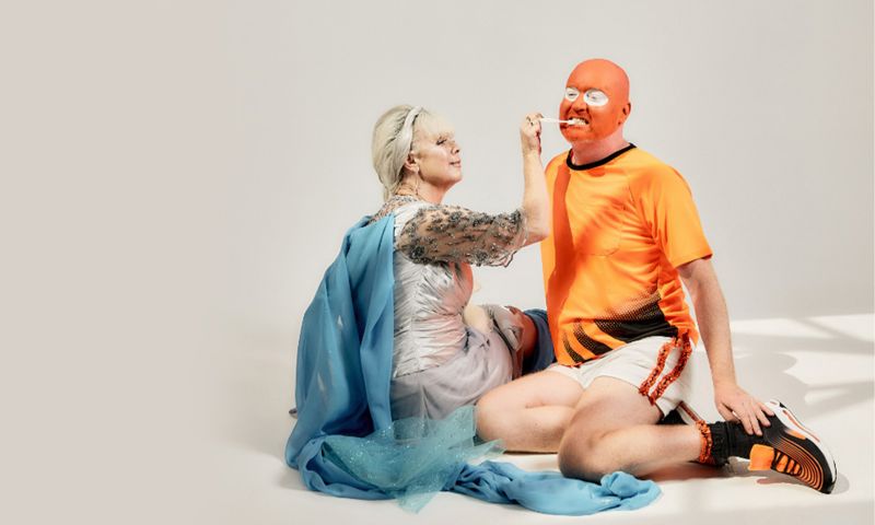 A person in a silver and blue dress brushes another person’s teeth. The person who is getting their teeth brushed is wearing orange face paint and an orange t-shirt with sport shorts.