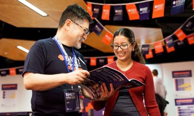 RMIT staff assisting a visitor by discussing details from an open day brochure inside a decorated venue with colourful RMIT banners hanging above. 