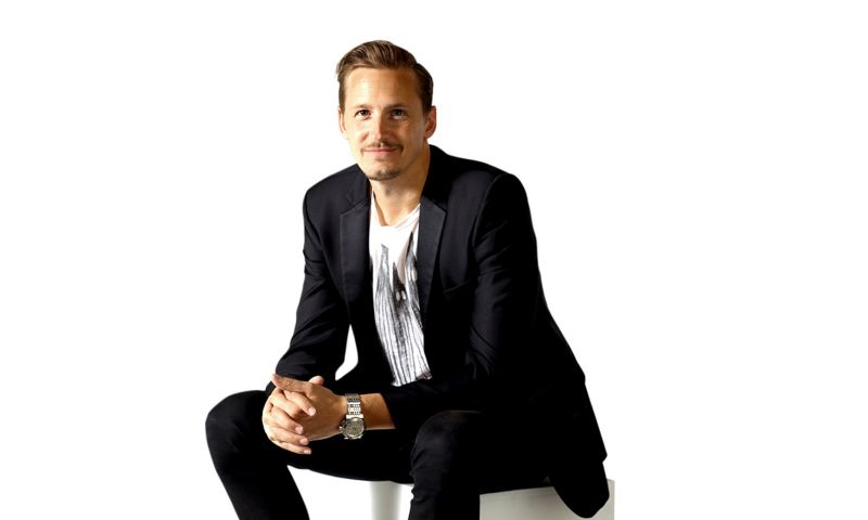 Pat Langton sitting in a studio for a photo wearing a dark grey suit and white t-shirt.