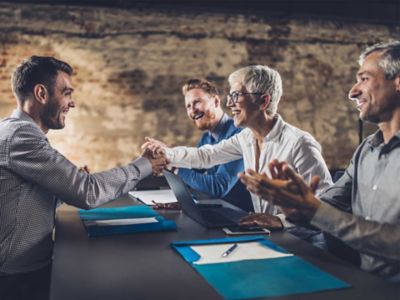 A candidate shakes hands with a panel interviewer. Two male panel interviewers clap in appreciation.