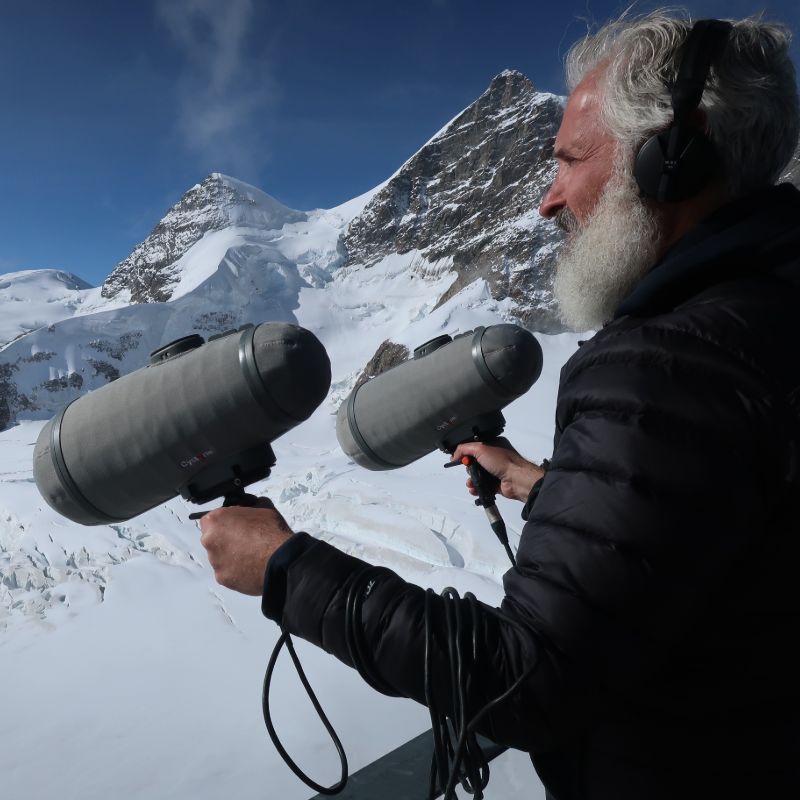 Photo of Philip Samartzis outdoors in a snowy environment. Rocky mountains in the background with snow covering it. Philip is standing on an elevated platform, with a railing in front of him. Philip is holding two large microphones in his hands, holding them a shoulder width apart. Philip has headphones on and is looking into the distance.