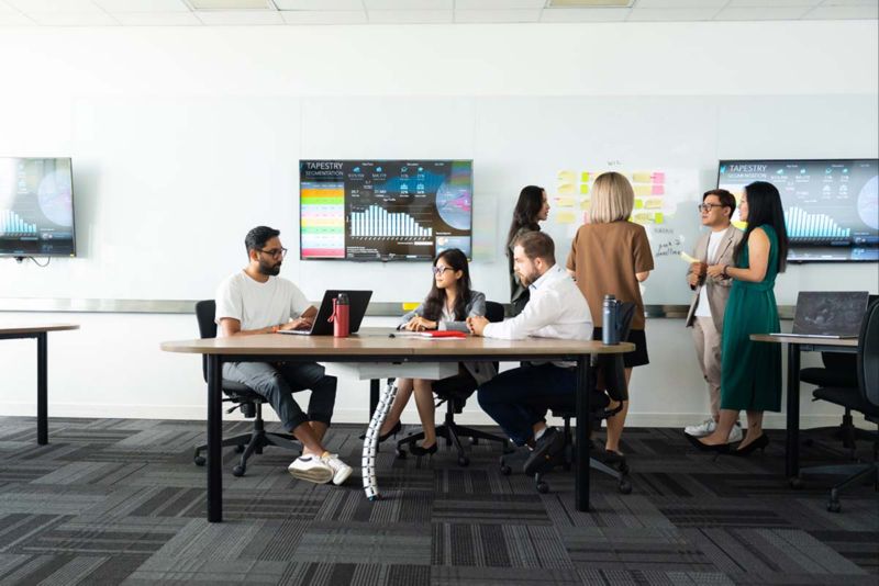 2 groups of multicultural postgraduate students in a classroom, holding separate discussions in breakout groups. Graphs flash from TV screens in the background.