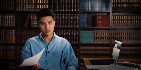  Male student sits at a desk in a formal University room holding papers in front of an open laptop.