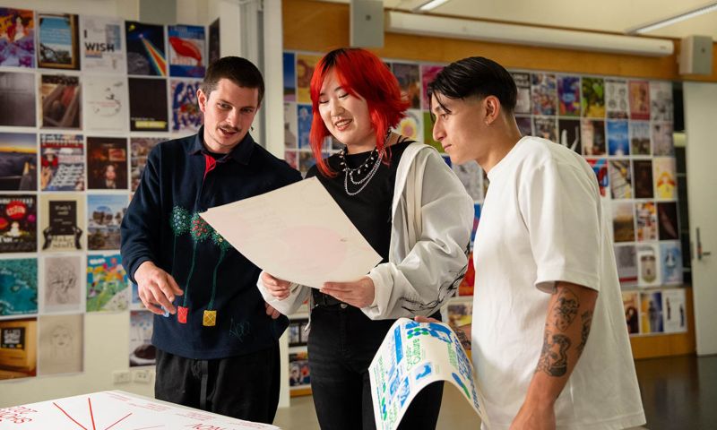 Three students in a classroom reading a paper together held by the middle student.
