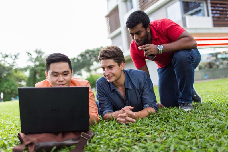 group of international students looking at laptop while lying on the grass