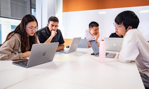 Five international students sitting on a desk with their laptops.