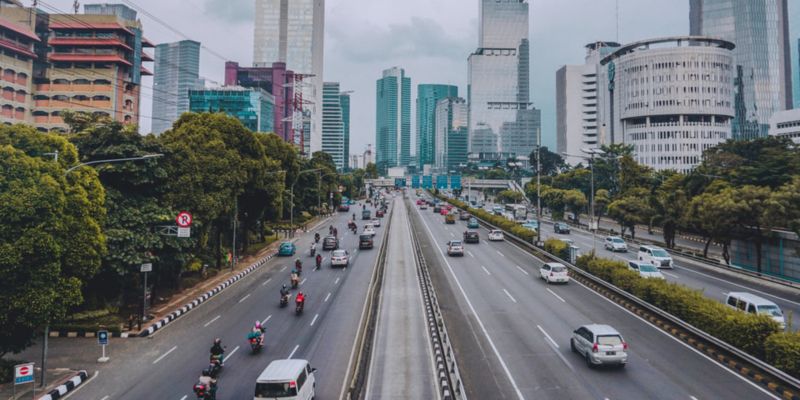 Photo of a busy highway. The city surrounds the highway with tall, mostly white buildings. There are trees and plants growing on the border of the highway.