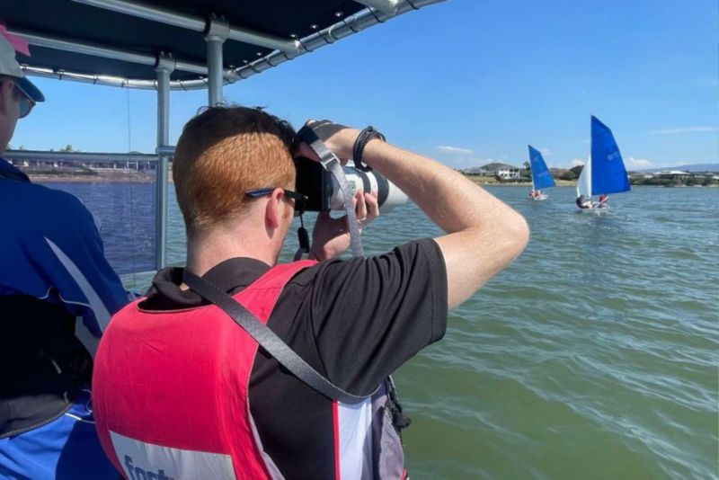 A person on a boat take photos of sailing ships in the distance. 
