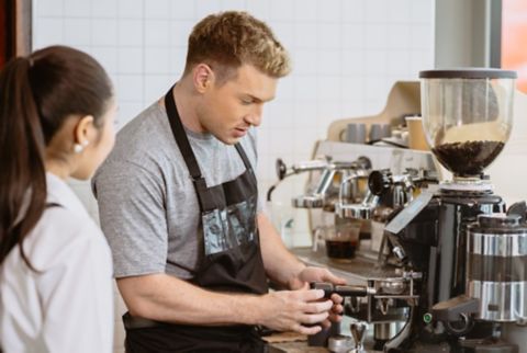 male barista uses coffee grinder in a cafe while a woman watches