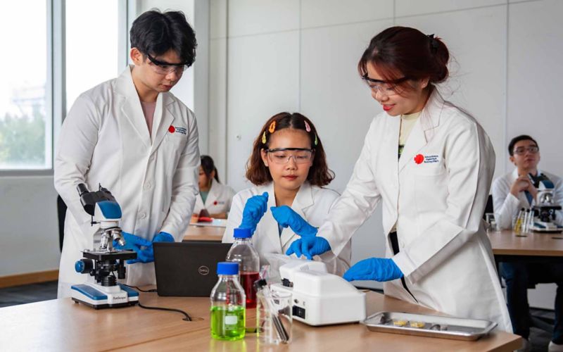 three students, 2 women and 1 man wearing RMIT white lab coats and safety glasses at a desk with a spectrometer and a microscope. A bottle of green fluid and a vial of dark brown fluid sitting in a beaker are in the front. To their side also sits a surgical plate with glass slides. 