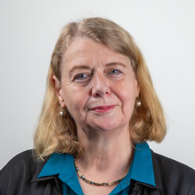 Sue is smiling towards the camera against a solid white background. Sue is wearing a blue shirt with a black cardigan.