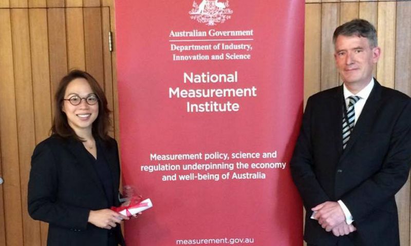 Suelynn Choy and another person standing on either side of a banner about the institute. Both are posing and smiling towards the camera.. Suelynn is holding a rolled up paper tied with a red bow in her hands.