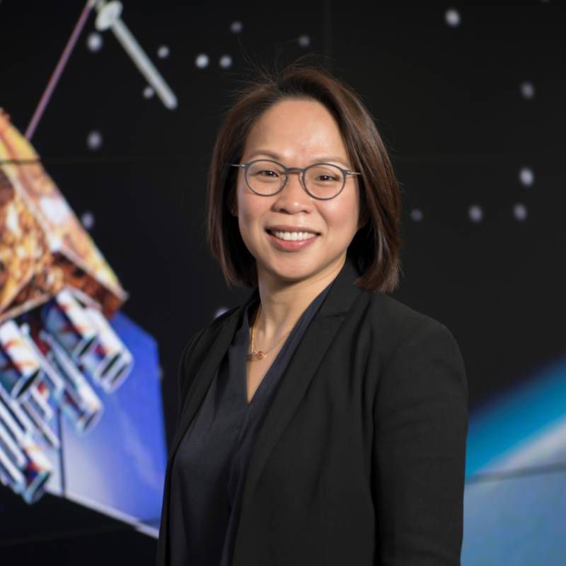 Profile photo of Suelynn Choy smiling towards the camera with an out of focus background of a banner illustrating a satellite in outer space