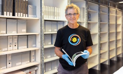 Tony Isaacson has short grey hair and glasses. He is wearing a black T-shirt and is standing in the archives holding a book.