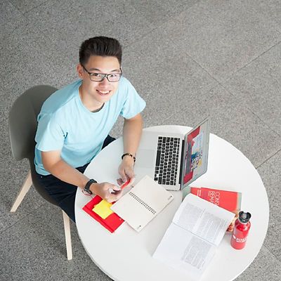 Vietnamese male student in a cyan t-shirt looking up from a desk with a laptop and study materials.