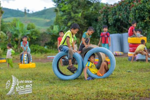 Children climb on and crawl through colourful recycled tyres on a grassy Wheel of dreams playground set against a backdrop of trees and hills.