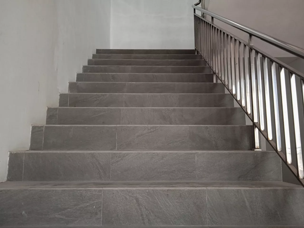 A minimalist staircase in an urban setting. The stairs, made of concrete, ascend in a straight line with a white handrail running alongside.