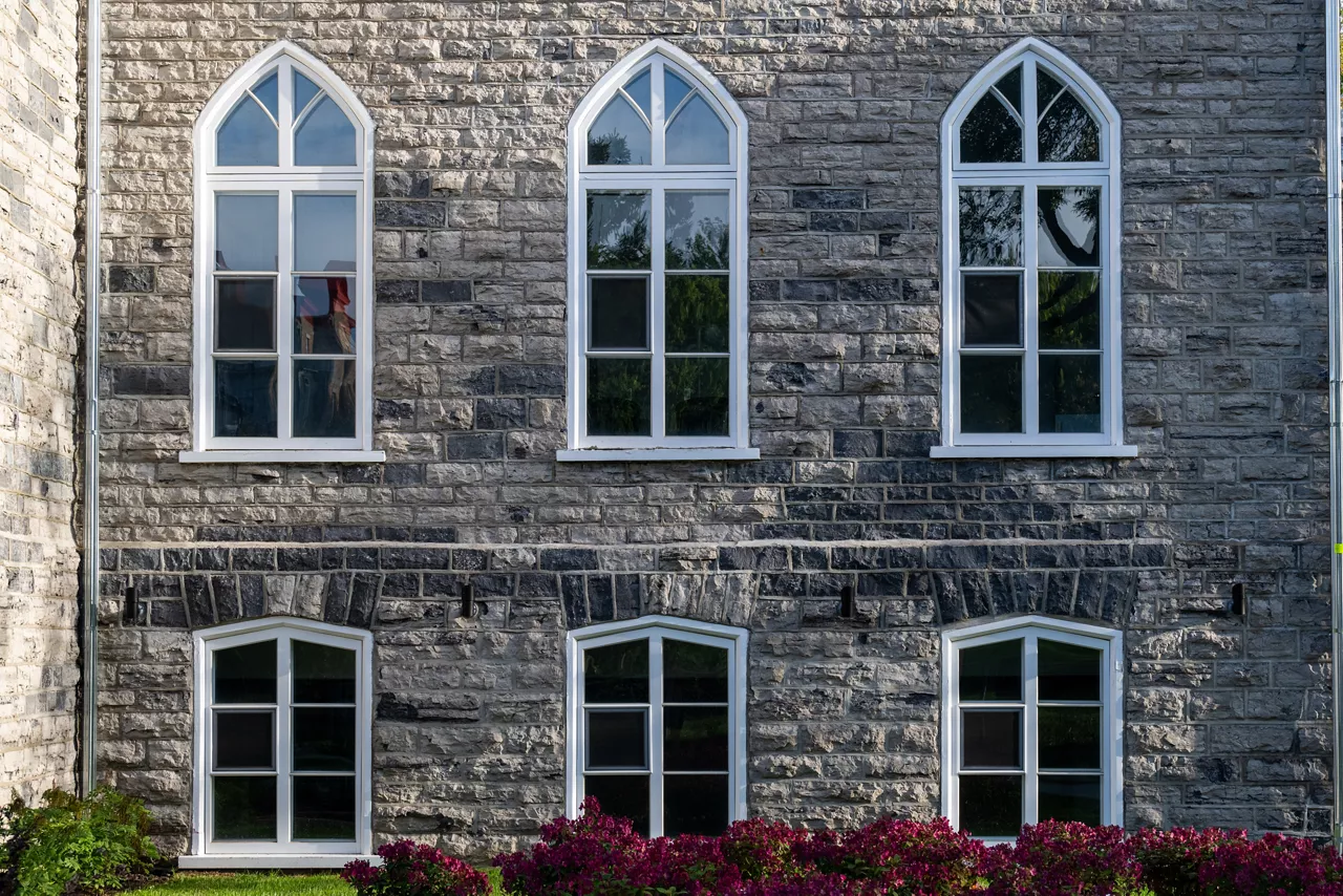 A historical concrete building, Gothic Revival style of architecture with multiple windows. Clean mason style with grey and beige bricks on the frame and has six clerestory cathedral glass windows.
