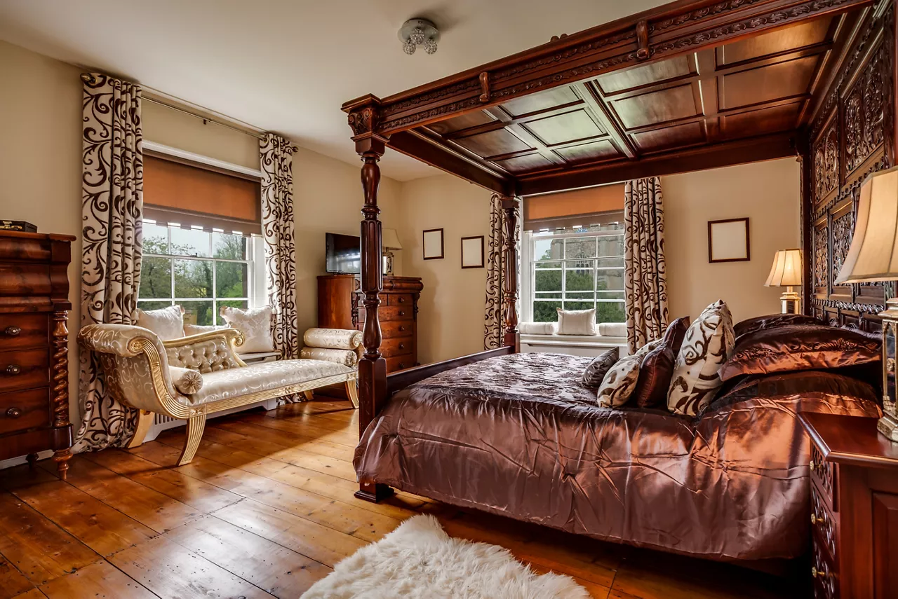 Furnished bedroom within former victorian rectory with ornate carved four poster bed and matching period sideboard drawers, chaise longe, side tables with lamps and exposed timber floor