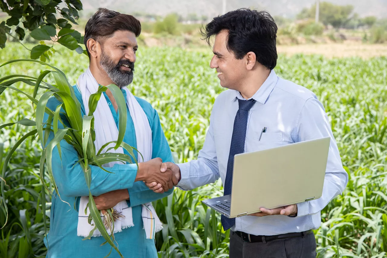 Happy Indian village farmer man and bank employee negotiating terms of agriculture loan in field, two men shaking hands in farm land standing with open laptop, skill India concept, Digital India.