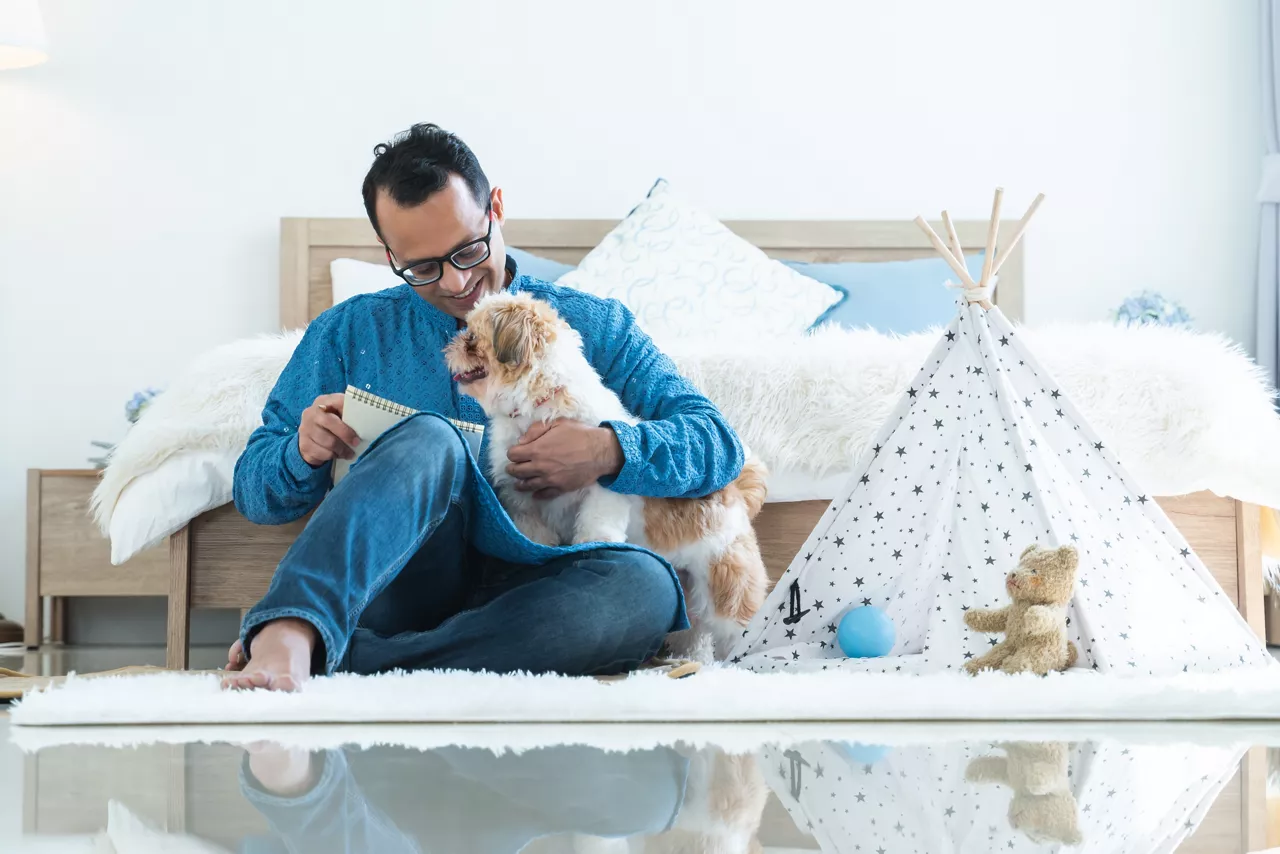 Indian middle aged man in traditional clothes working sitting on floor, relaxing playing with shih tzu dog puppy in bedroom at home. Pet friendly residence, lifestyle, pet and human bonding concept