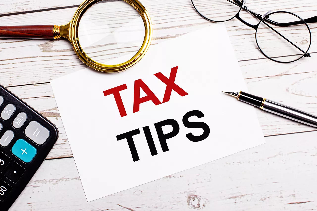 A workplace with a pen, glasses, a calculator, a magnifying glass and a white sheet of paper with the text of the TAX TIPS on a white wooden table. Business concept.