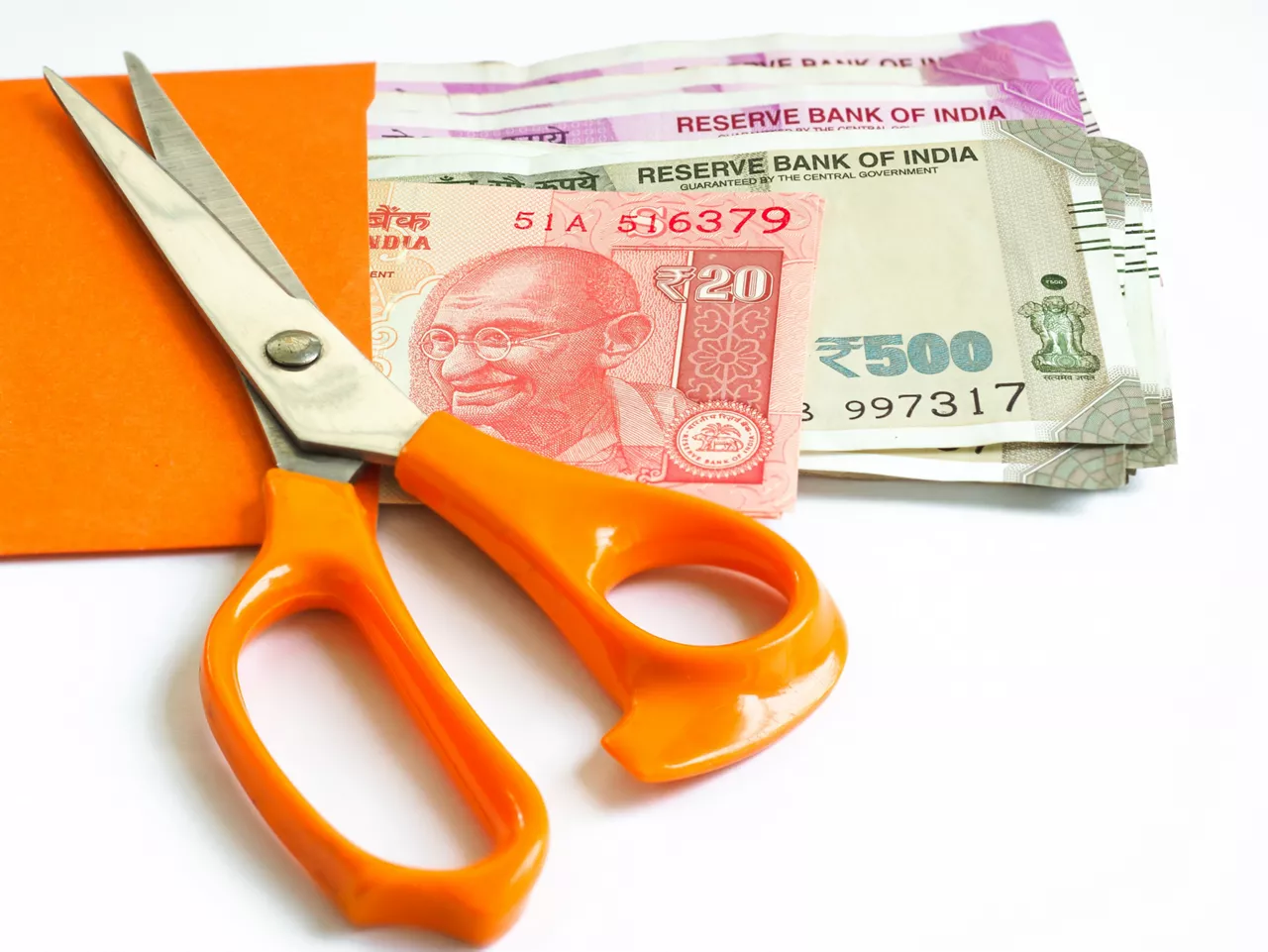 The stainless steel scissors with orange plastic handles is on top of the orange envelope which contain Indian Rupees on the white background