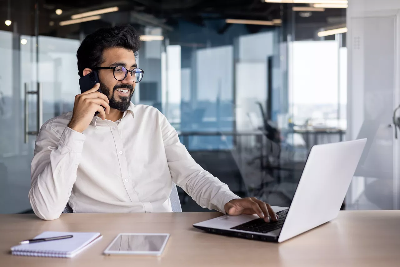A smiling young Indian man is sitting at a desk in the office, using a laptop and talking on the phone.