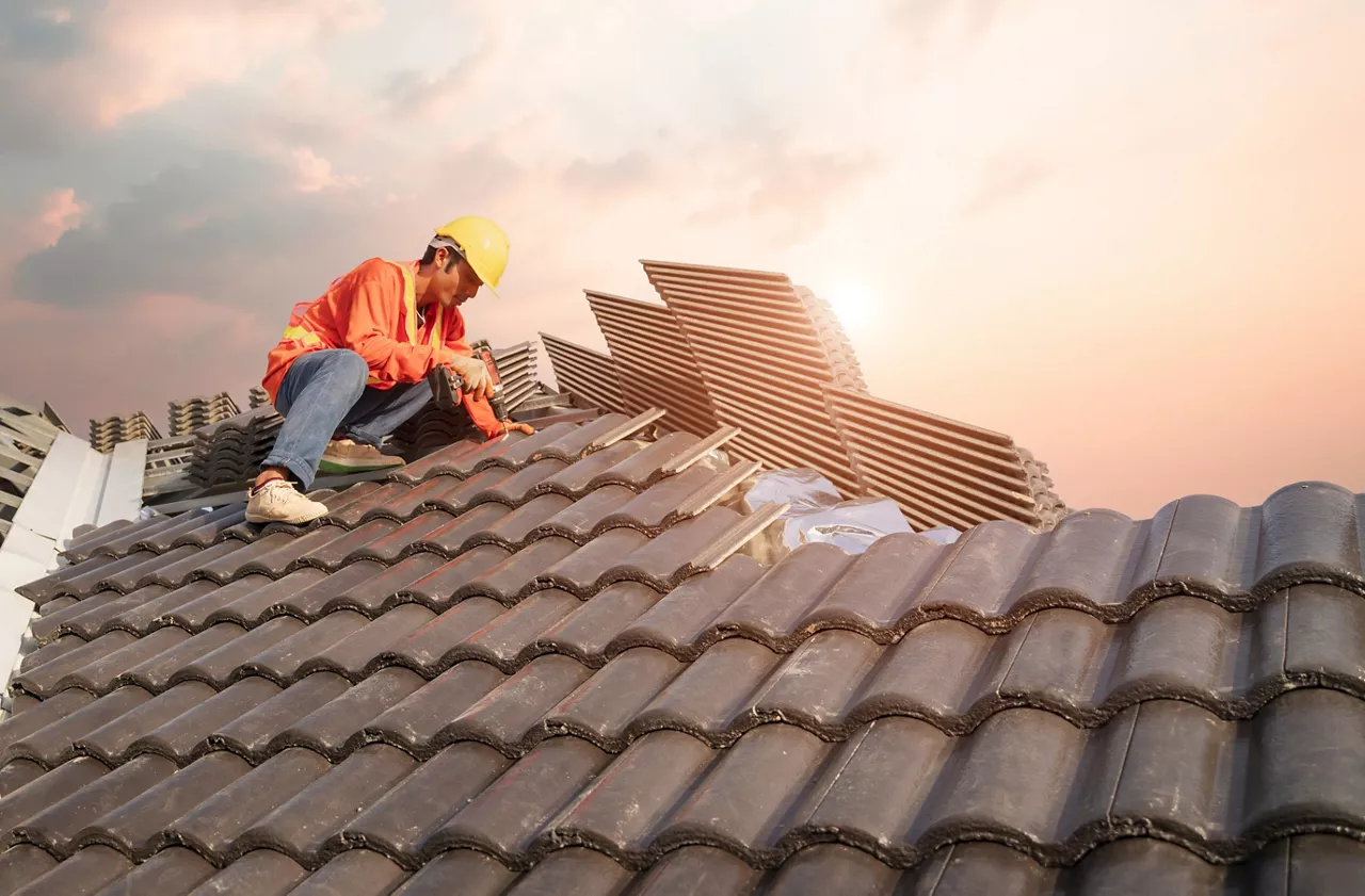 A male roofer uses a cordless electric battery drill to fire screws to install concrete roofing slabs or c-packs on home structure construction