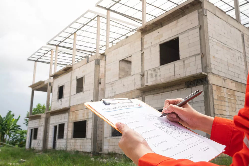 The architect or project engineer is checking on house building quality checklist for inspecting the building quality, with house structure as background. Industrial working scene.