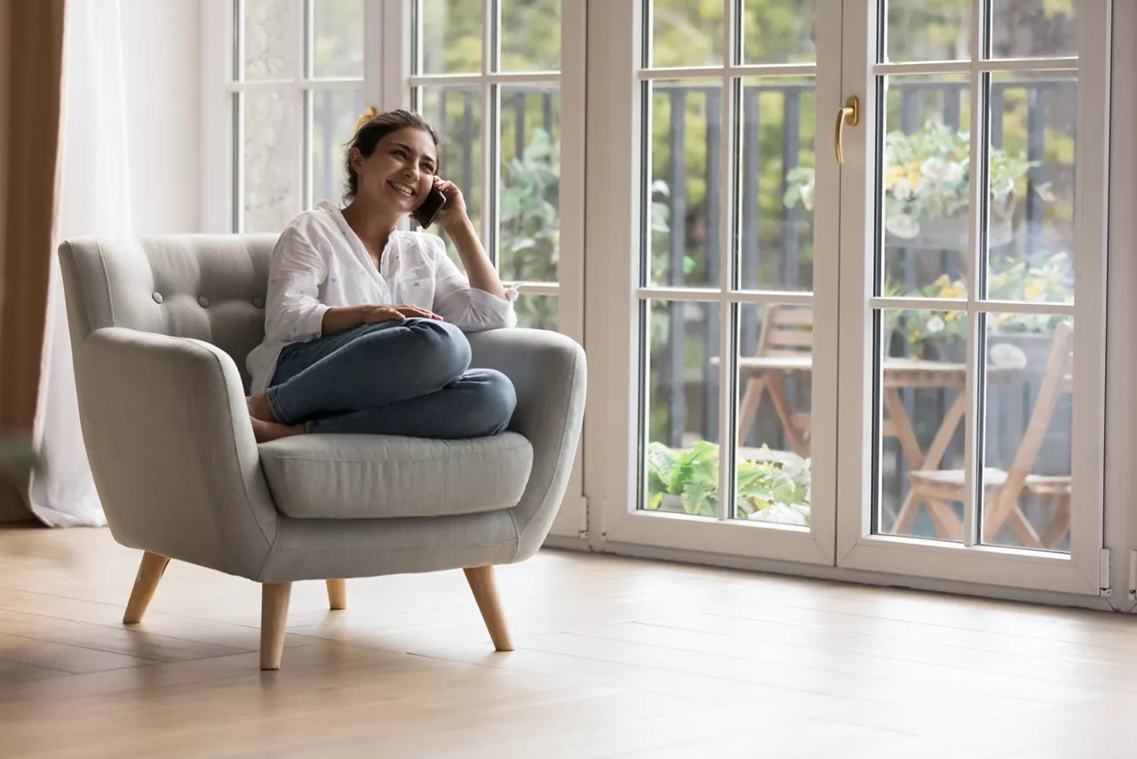 Cheerful happy millennial Indian girl speaking on mobile phone, sitting in single armchair at panoramic terrace window, enjoying funny talk, leisure, communication, smiling, laughing. Copy space