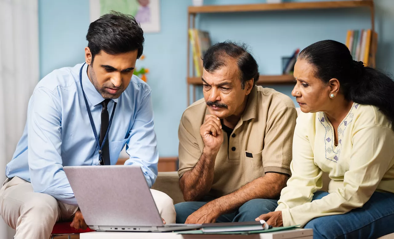 Indian banker explaining about insurance policy or savings scheme on laptop to senior couple while sitting on sofa at home - concept of banking support, and bonding