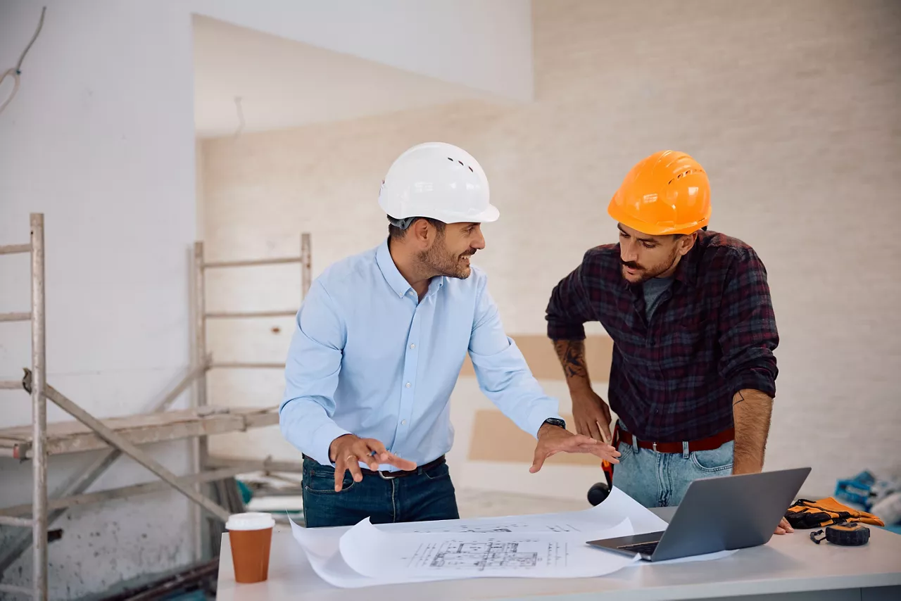 Happy architect talking to construction worker while analyzing blueprints during house remodeling. 