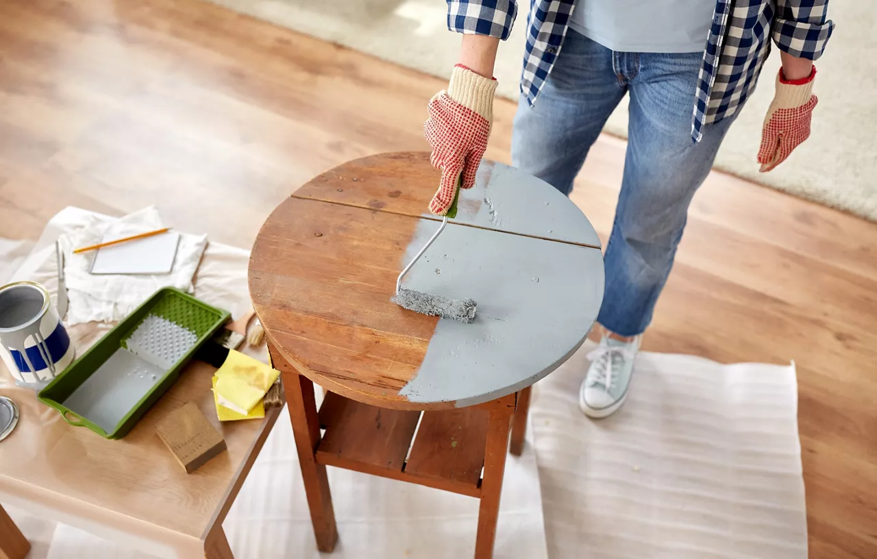 furniture renovation, diy and home improvement concept - close up of woman in gloves with paint roller painting old wooden table in grey color