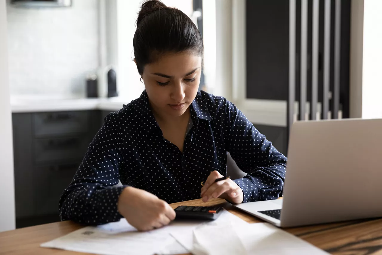 Home office. Concentrated young lady of indian ethnicity sitting at desk in modern studio apartment engaged in paperwork. Hindu woman counting preparing to pay taxes bills using computer calculator