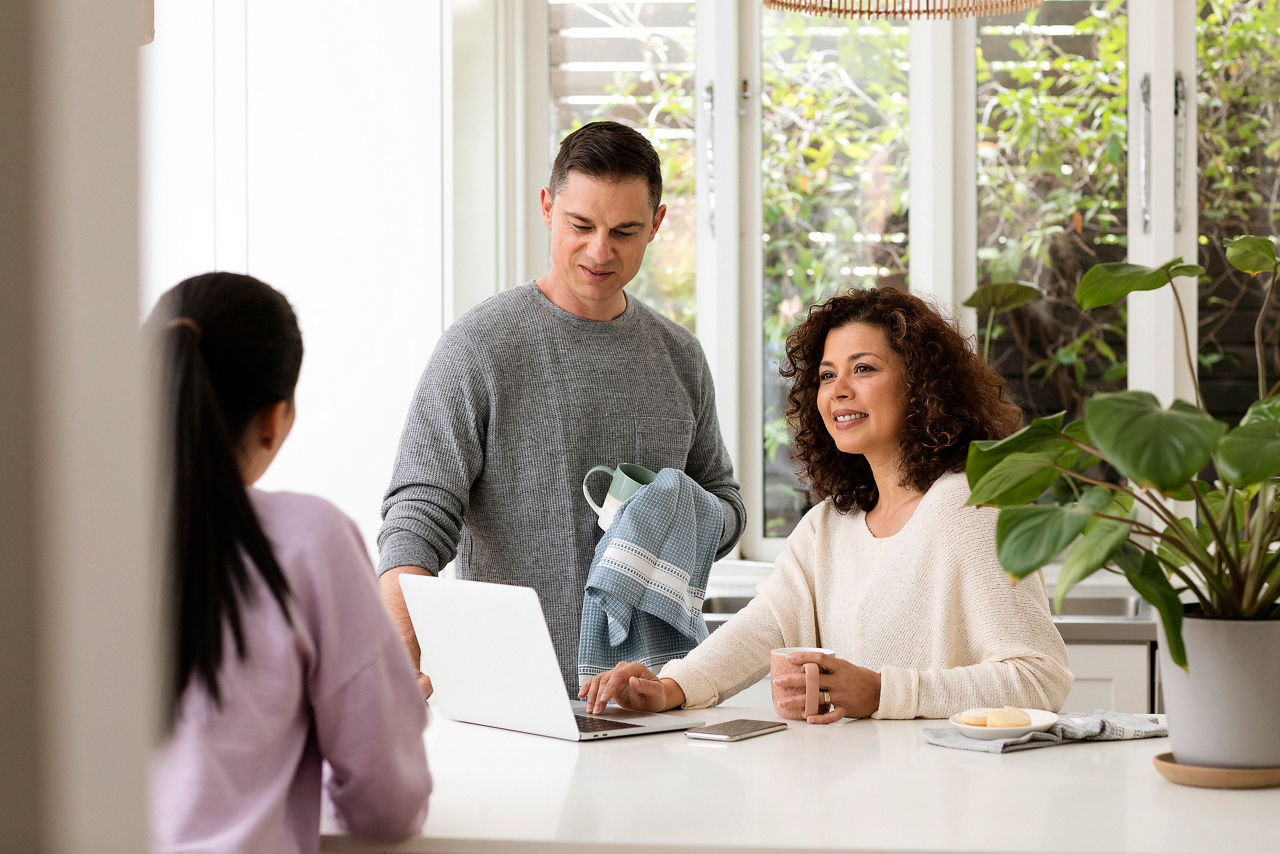 Family at the kitchen bench, man drying cup, woman on laptop chatting with girl
