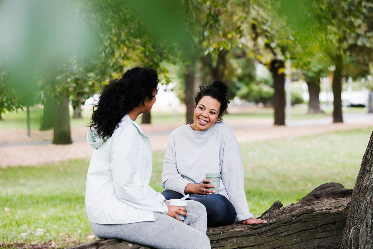 Two ladies sitting on a log in the park chatting with coffee in hand