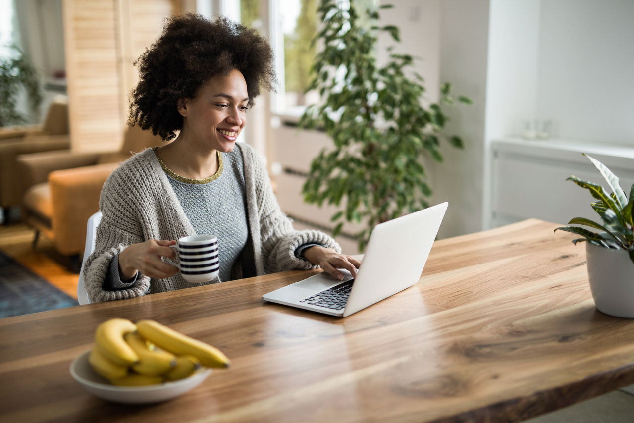 Smiling black woman relaxing at home while using computer during her coffee time.