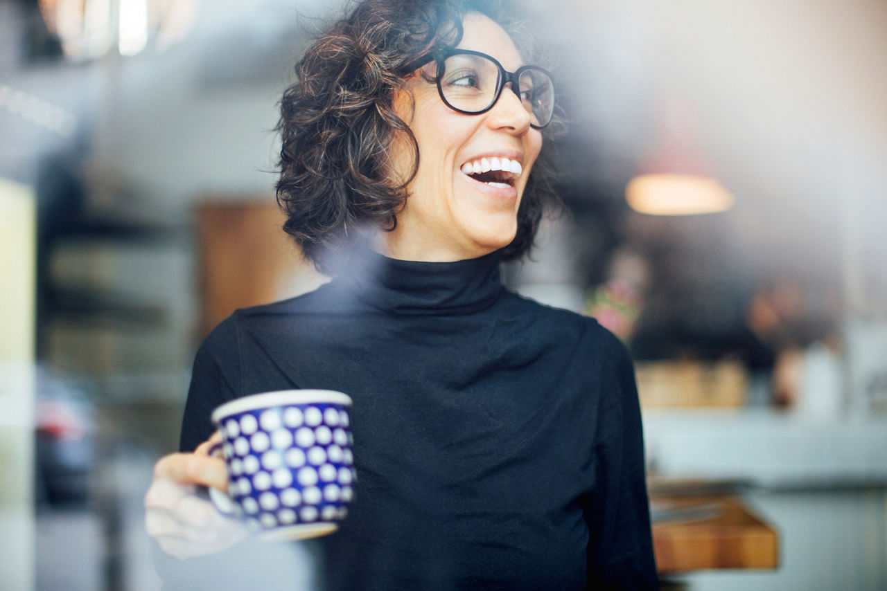 Mature businesswoman laughing while coffee at coffee shop. Mid adult female sitting inside cafe with a coffee in hand.