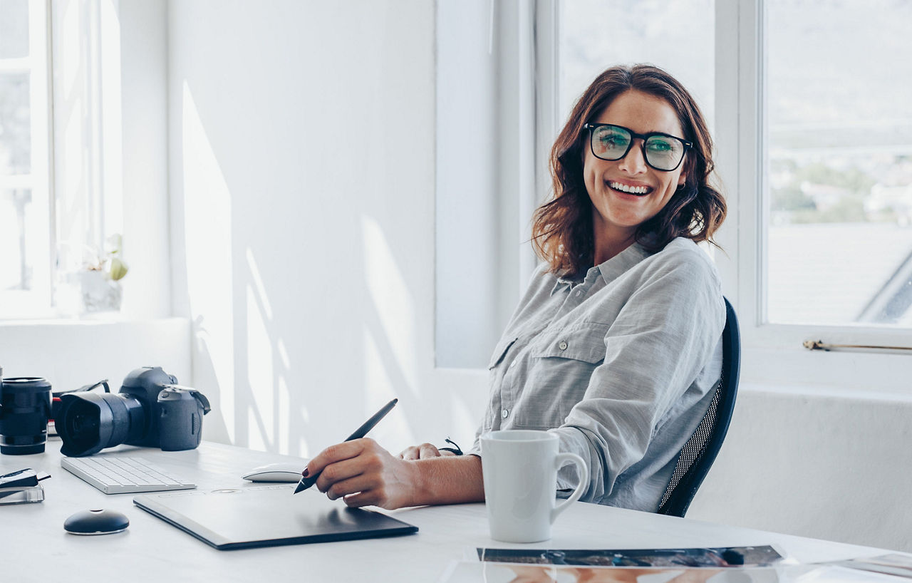 Professional photographer sitting at her office desk looking away and smiling. Woman in office with digital graphic tablet and drawing pen.