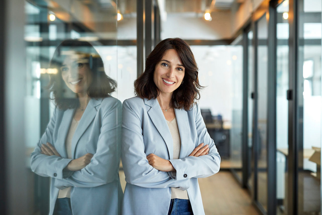 Portrait of confident businesswoman with arms crossed. Smiling mid adult female professional is wearing smart casuals. She is leaning on glass wall in corridor at creative office.