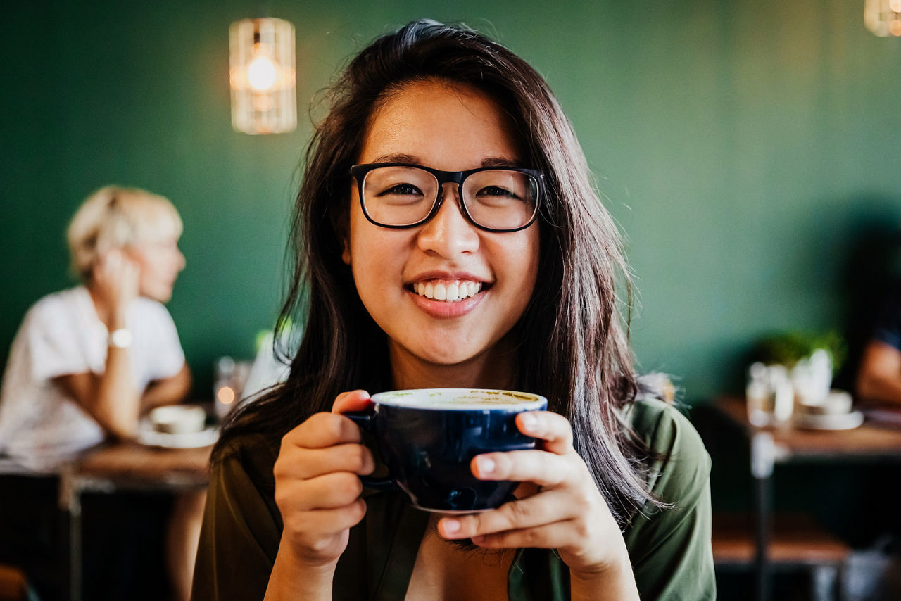 A portrait of a young Asian woman with glasses, smiling while drinking coffee in her favorite cafe.