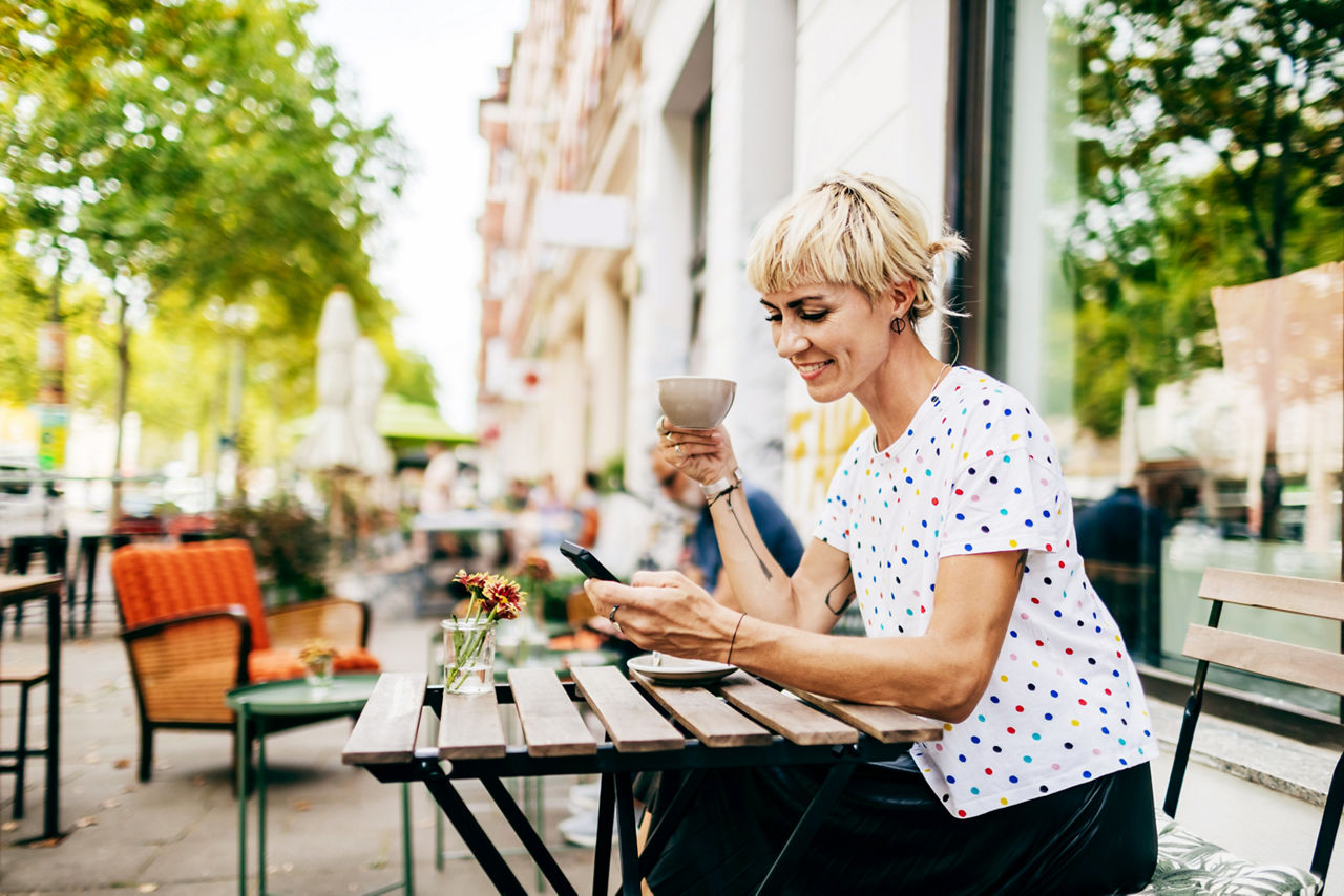 A woman sitting at a table outside a cafe looking at her smartphone while drinking a cup of coffee.